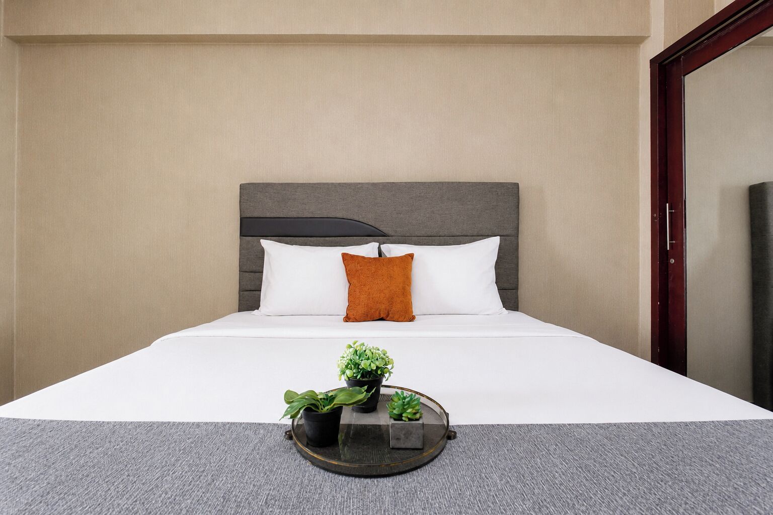 Una camera d'albergo con un letto, una testata, un comodino con vassoio e una pianta, e uno specchio.