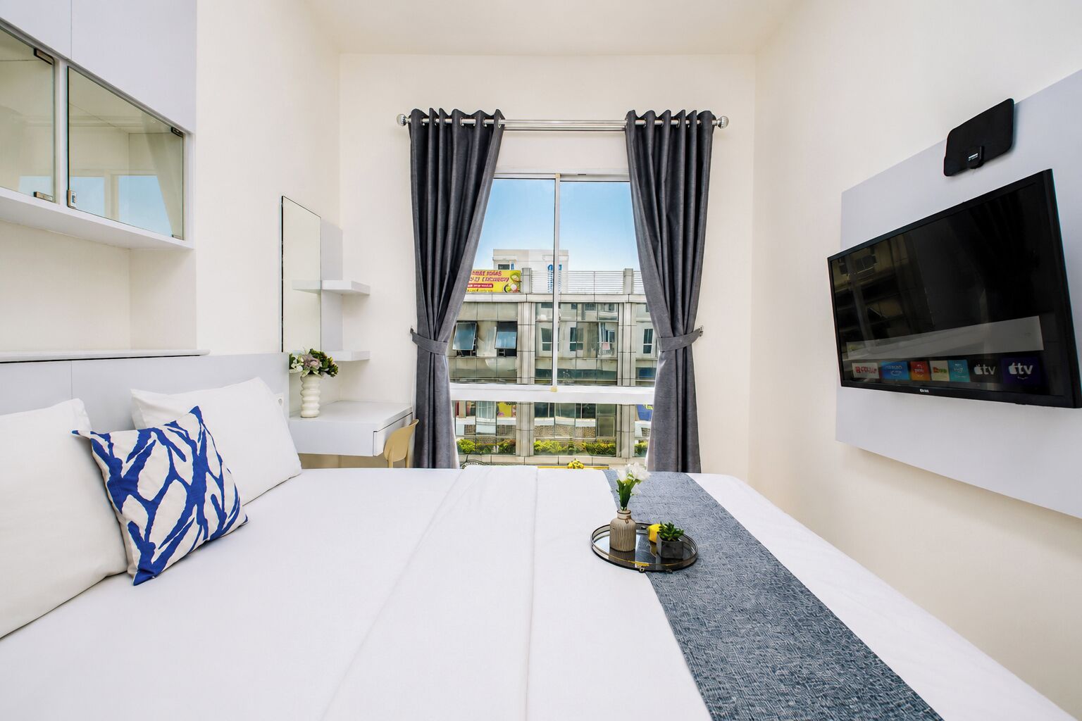 A hotel room with a bed, a TV, a window with curtains, and a view of buildings outside.