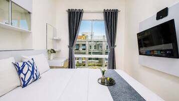 A hotel room with a bed, a TV, a window with curtains, and a view of buildings outside.