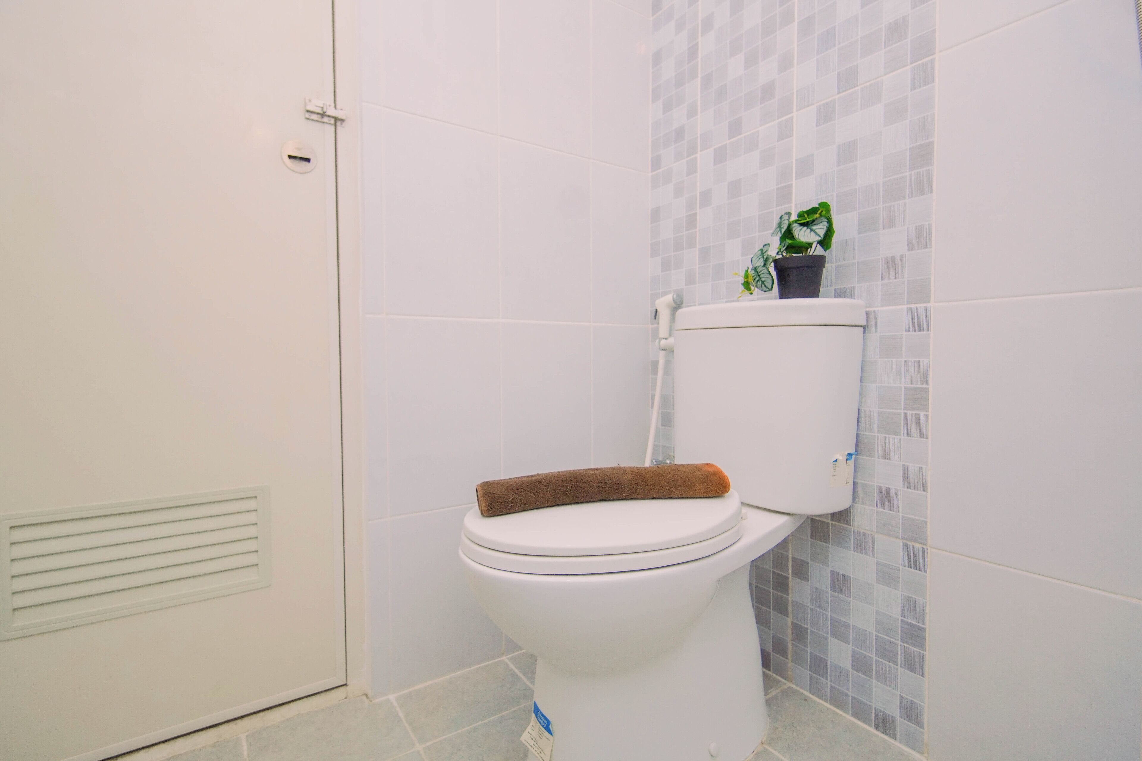 Eine weiße Toilette mit einem braunen Handtuch auf dem Sitz, einer Topfpflanze auf dem Tank und einer gefliesten Wand mit Rastermuster.