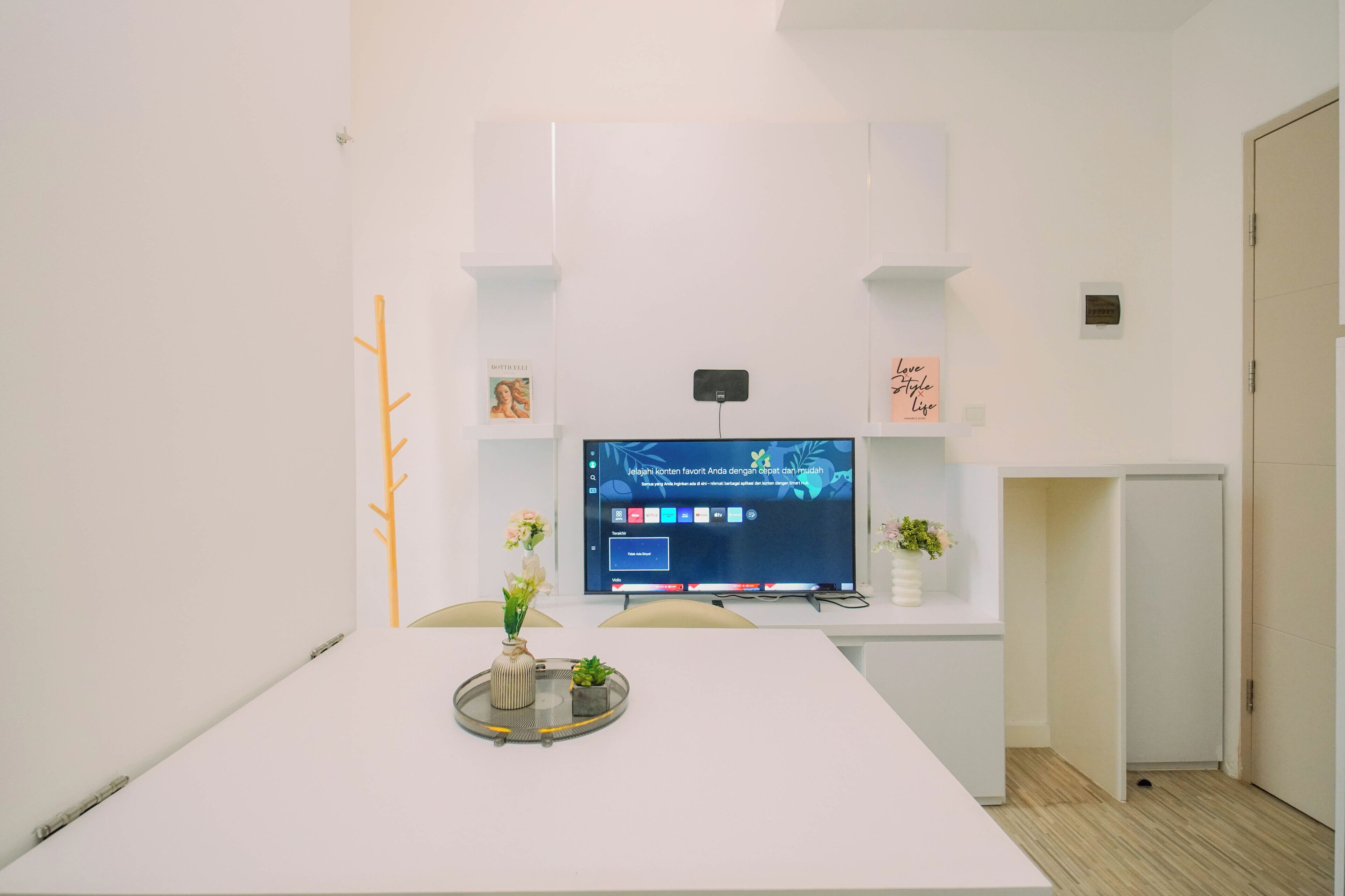 A modern, minimalist office space with a large flat-screen TV mounted on a white wall, a white table with a vase of flowers, and a small potted plant.