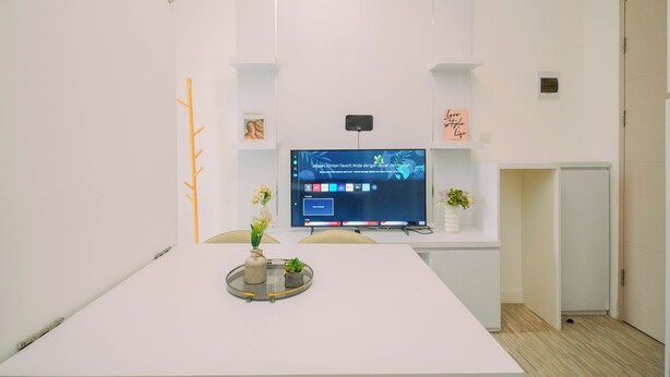 A modern, minimalist office space with a large flat-screen TV mounted on a white wall, a white table with a vase of flowers, and a small potted plant. - Warm Minimalist 2Br At Apartment Tokyo Riverside Pik 2 (Jakarta)
