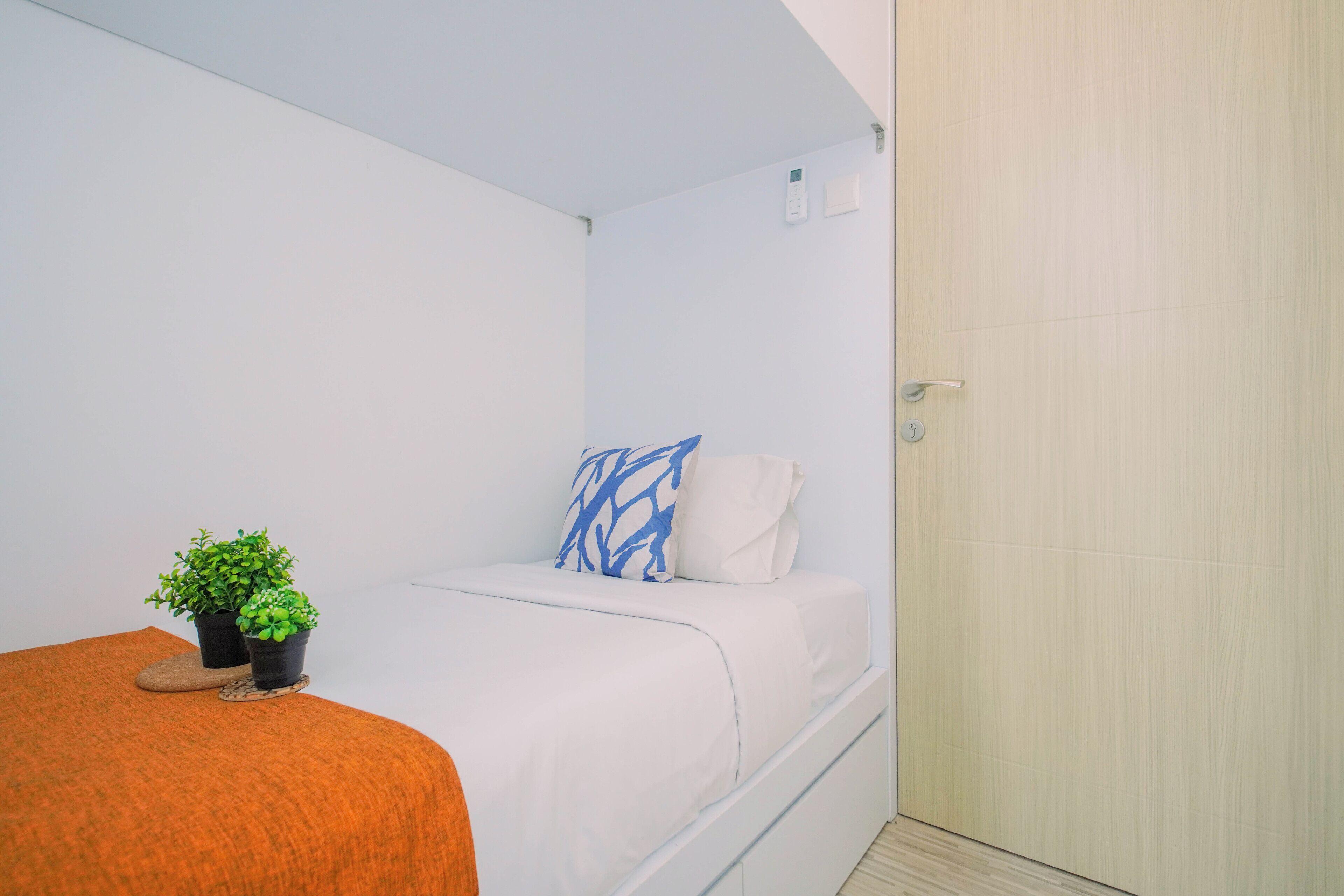 Een kleine kamer met een enkel bed, een wit bed, een blauw-wit kussen, een kleine potplant en een houten deur.