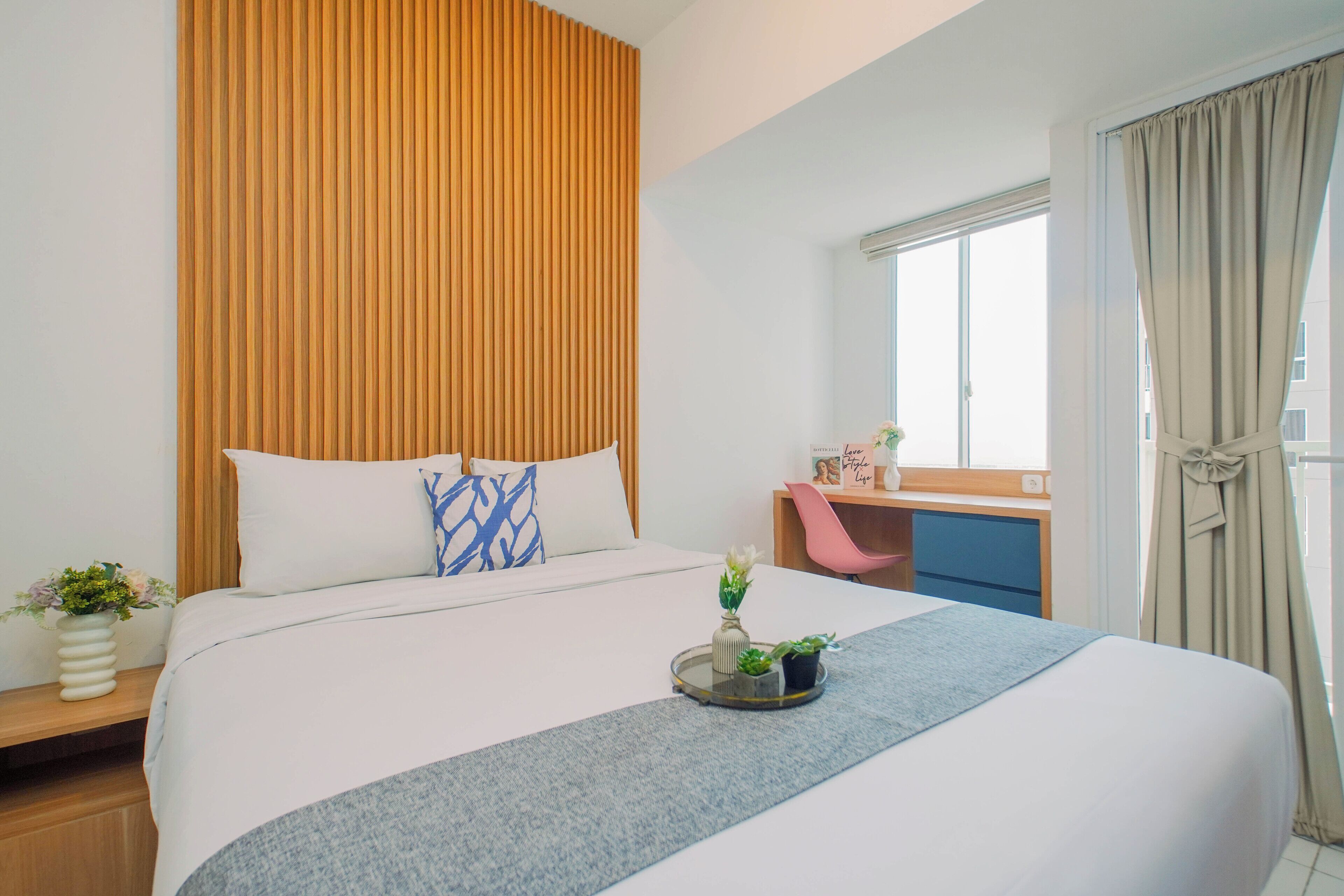 Hotellihuone, jossa on sänky, työpöytä, tuoli, ikkuna verhoilla ja pieni kasvi yöpöydällä.