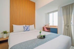 A hotel room with a bed, a desk, a chair, a window with curtains, and a small plant on the bedside table.