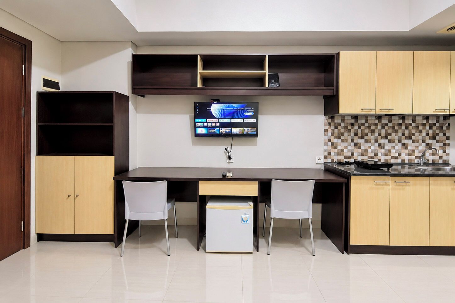 A compact kitchen with a built-in TV, two white chairs, and a small refrigerator.