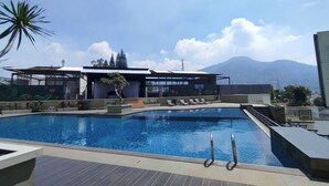 A swimming pool with lounge chairs, a covered seating area, and a mountain view in the background. - Urban Cozy Studio At Apartment Skyland City Jatinangor (Sumedang)