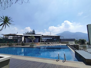 A swimming pool with lounge chairs, a covered seating area, and a mountain view in the background. - Urban Cozy Studio At Apartment Skyland City Jatinangor (Sumedang)