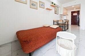 Une pièce avec un lit simple, une petite table et une chaise. On y trouve des tableaux encadrés au mur, un coin cuisine au fond de la pièce et une porte.
