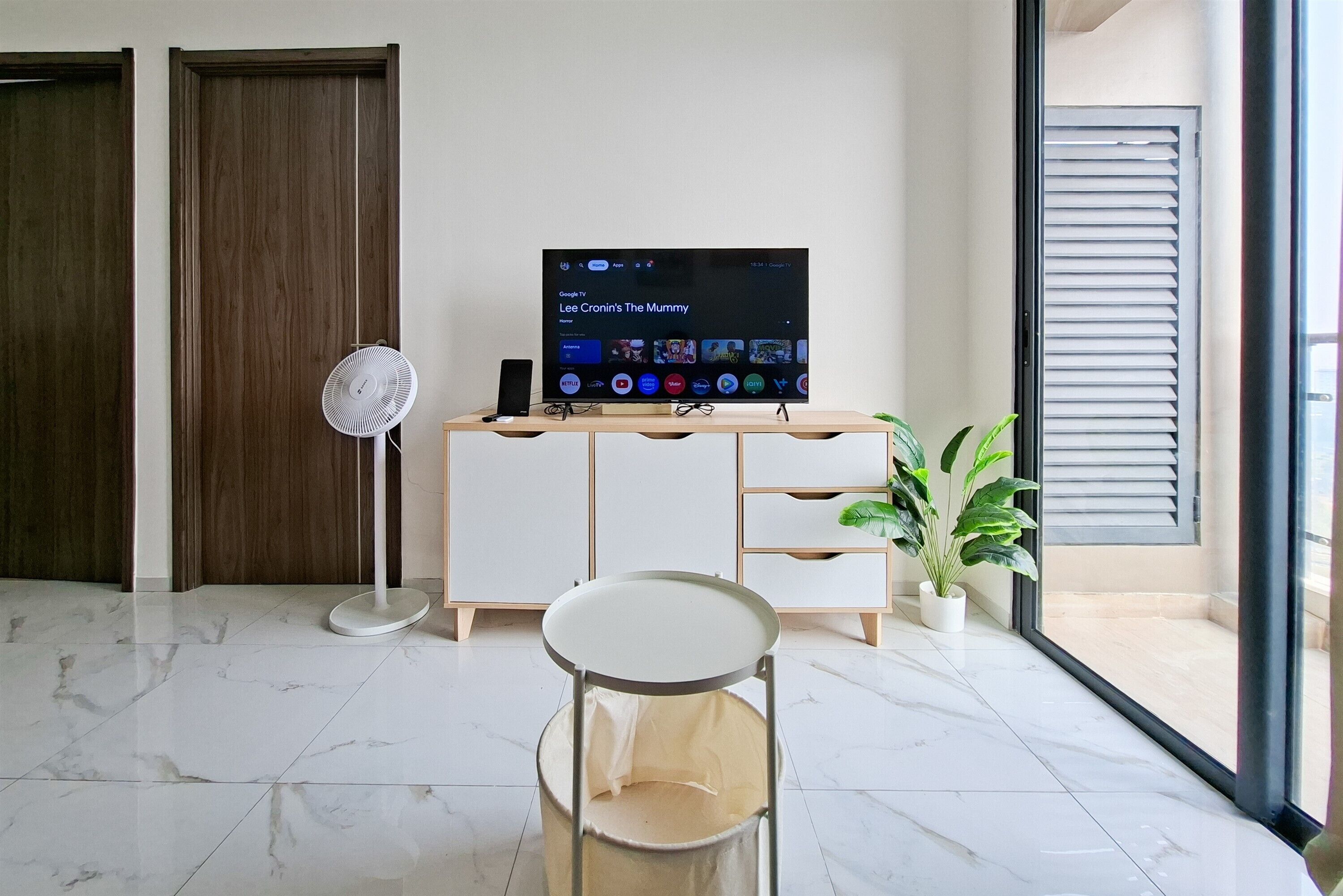 A modern living room with a flat-screen TV, a standing fan, a white cabinet, a round table, and a potted plant.