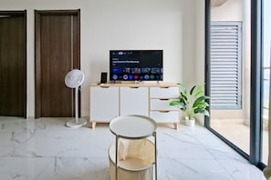 A modern living room with a flat-screen TV, a standing fan, a white cabinet, a round table, and a potted plant. - Beautifully Designed 2Br At Apartment Sky House Bsd (Pagedangan)