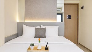 A hotel room with a bed, a bedside table with a vase and a plant, and a wooden door. - Best Deal Studio At Apartment Meikarta District 2 (Cikarang)
