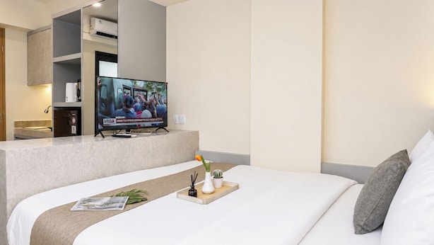 A hotel room with a bed, a TV on a stand, a small table with a tray, and a potted plant. - Best Deal Studio At Apartment Meikarta District 2 (Cikarang)