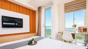 A hotel room with a bed, a desk with a chair, a TV mounted on the wall, and a window with a view. - Warm Studio Room With City View At Apartment Tokyo Riverside Pik 2 (Teluknaga)