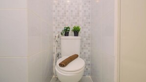 A white toilet with a towel on top, two potted plants on the tank, and a tiled wall with a grid pattern. - Warm Studio Room With City View At Apartment Tokyo Riverside Pik 2 (Teluknaga)
