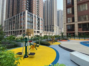 A playground with exercise equipment surrounded by greenery and buildings in the background. - Homey And Warm Studio Room At Apartment Meikarta District 1 (Cikarang)