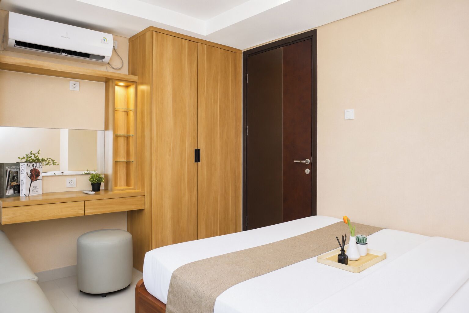 A hotel room with a bed, a wooden wardrobe, a small bedside table, a mirror, and an air conditioning unit.