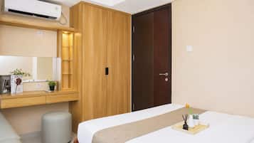 A hotel room with a bed, a wooden wardrobe, a small bedside table, a mirror, and an air conditioning unit.