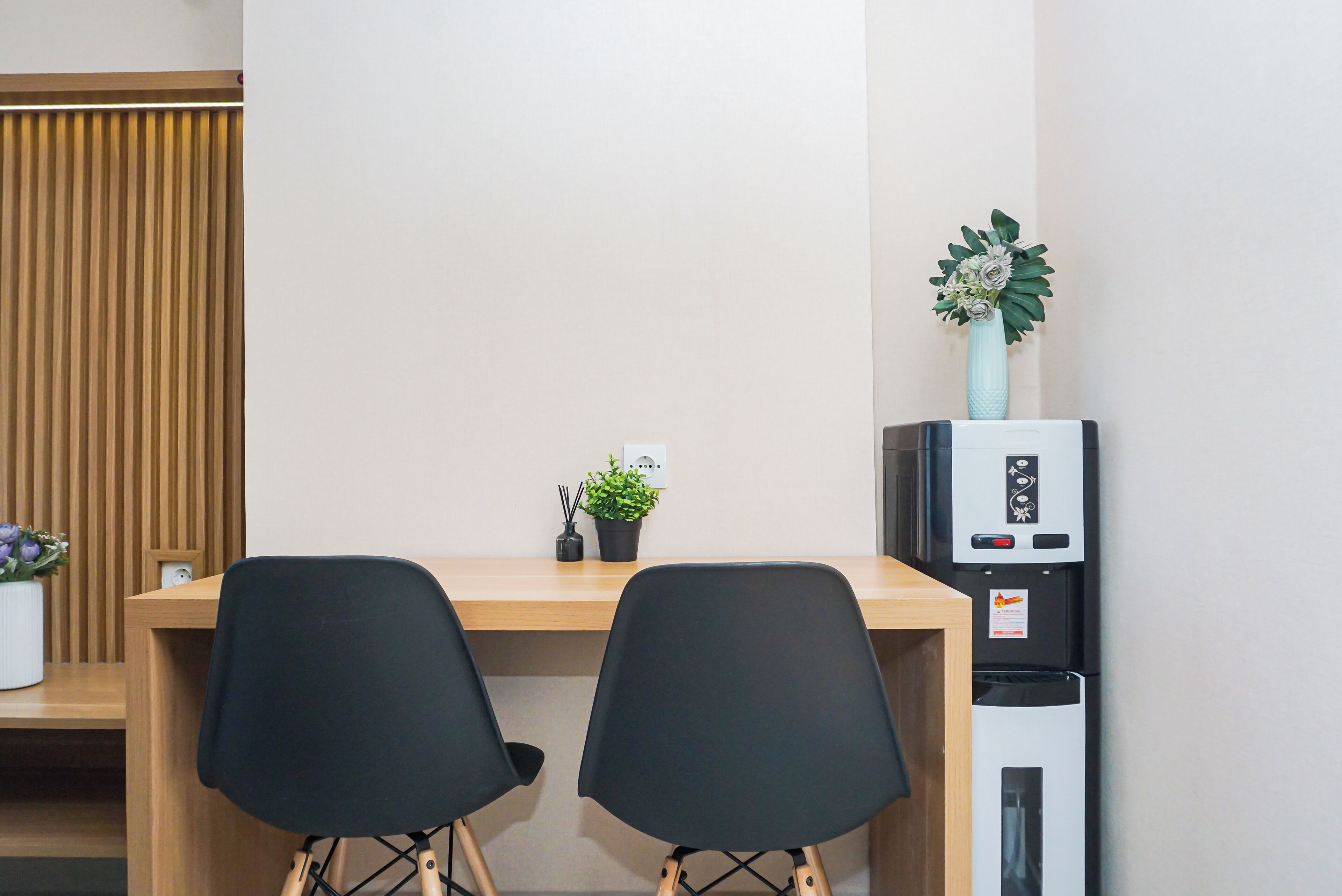 Quarto com escrivaninha de madeira, duas cadeiras pretas, um vaso com planta pequena, dispensador de água e quadro branco.