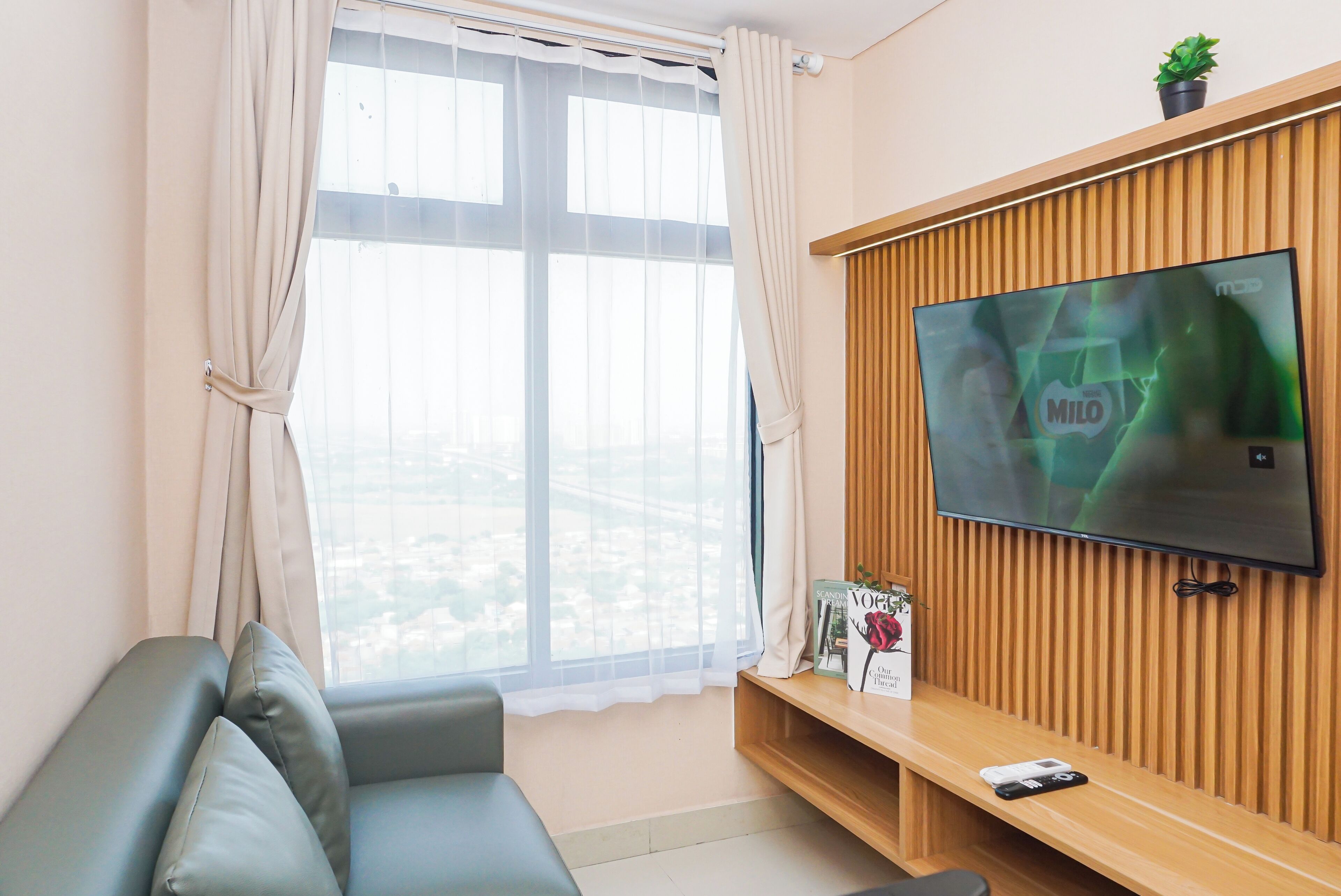 A modern living room with a large window, a flat-screen TV mounted on a wooden panel wall, a grey sofa, and a small table with a remote control.