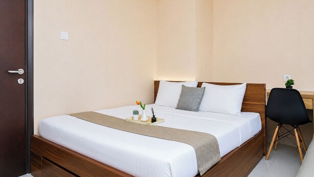 A hotel room with a bed, bedside table, chair, and a small desk with a plant. - Modern Look 2Br Apartment Pollux Chadstone (Cikarang)