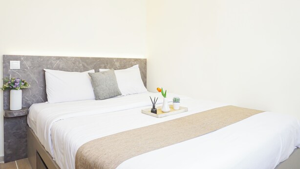 Cama bem arrumada com lençóis brancos e manta bege, mesa de cabeceira com um vaso de flores e bandeja com objetos decorativos.
