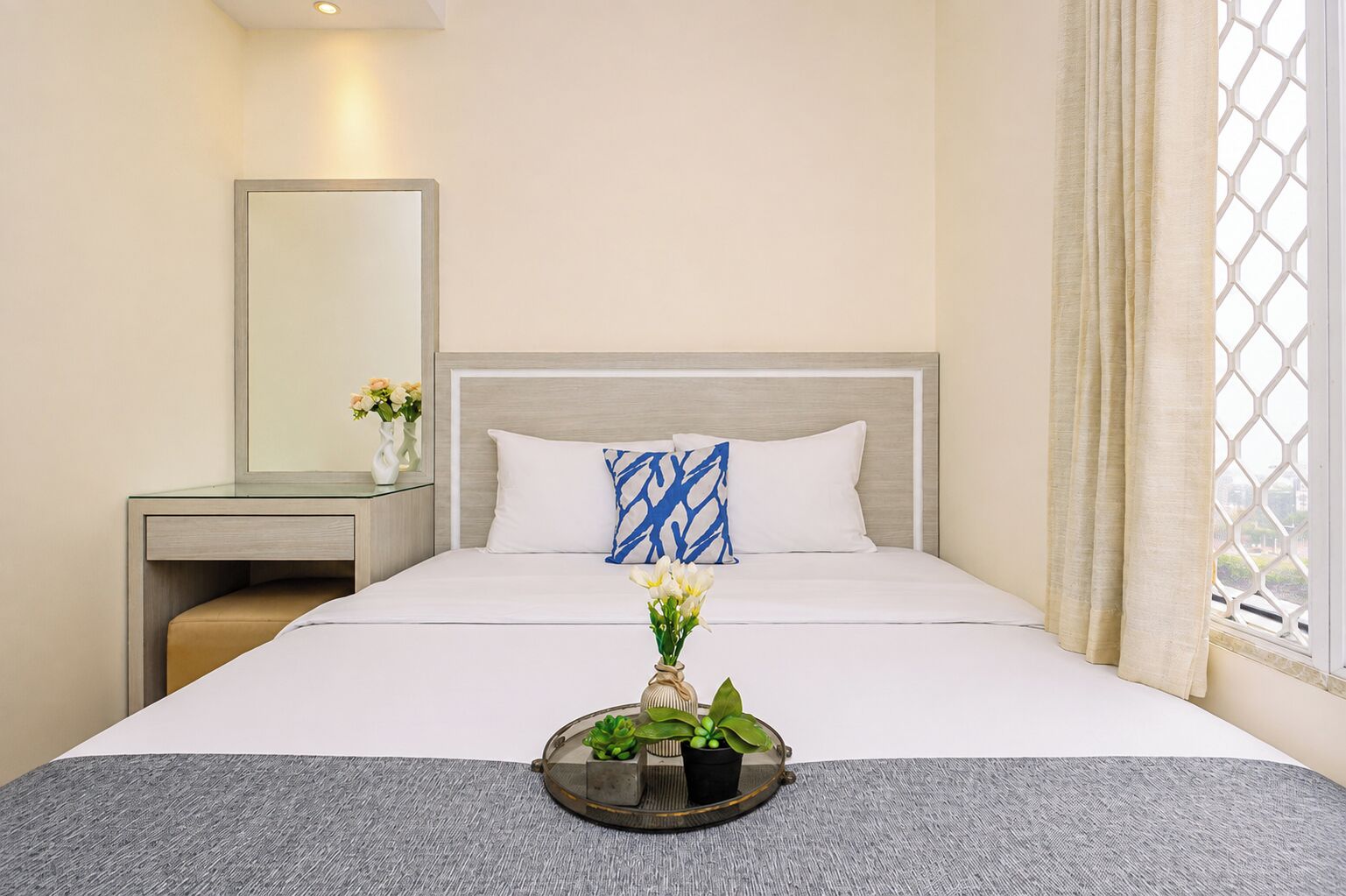 Een hotelkamer met een bed, nachtkastje, spiegel, raam met gordijnen, en een dienblad met bloemen en een plant.