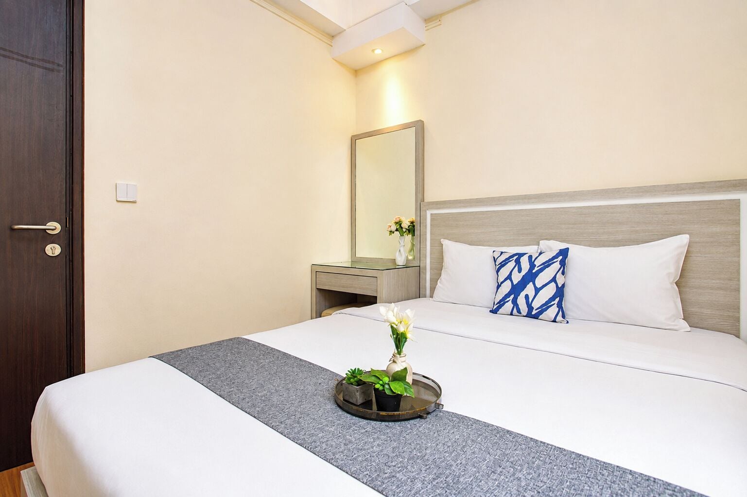 A hotel room with a bed, a bedside table, a mirror, and a vase with flowers.