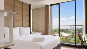 A hotel room with a large bed, a nightstand, a chair, and a view of a cityscape through large windows.