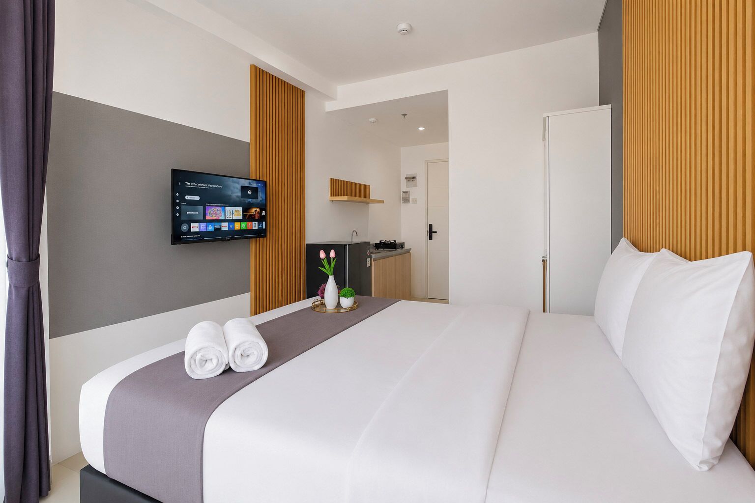 A hotel room with a bed, a flat-screen TV mounted on the wall, a small kitchenette, and a door leading to another room.
