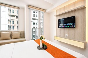 Habitación de hotel con cama, televisión montada en la pared, ventana con vistas a edificios y una mesita con un florero.