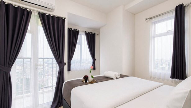 A hotel room with a bed, a bedside table with a vase and a tray, and a window with curtains. - Warm And Stylish 1Br At Apartment Kyo Society (Surabaya)