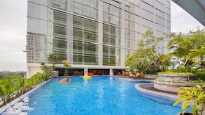 A swimming pool with clear blue water, surrounded by greenery and a modern building with large windows. - Enjoyable Studio At Apartment Mataram City Palagan Yogyakarta (Ngaglik)