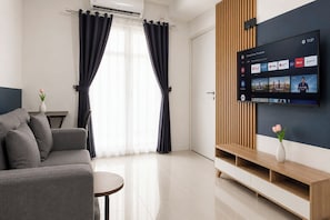 A modern living room with a grey sofa, a small round table, a TV mounted on a wall, and a wooden TV stand. - Stylish Bright 1Br At Apartment Kyo Society (Surabaya)