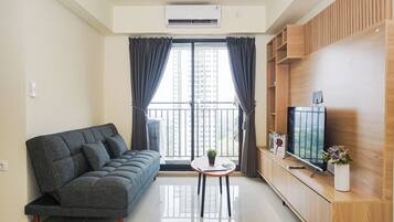 Una sala de estar moderna con un sofá gris, una mesita con una planta, un televisor de pantalla plana sobre un mueble de madera y un balcón con vistas a edificios altos.