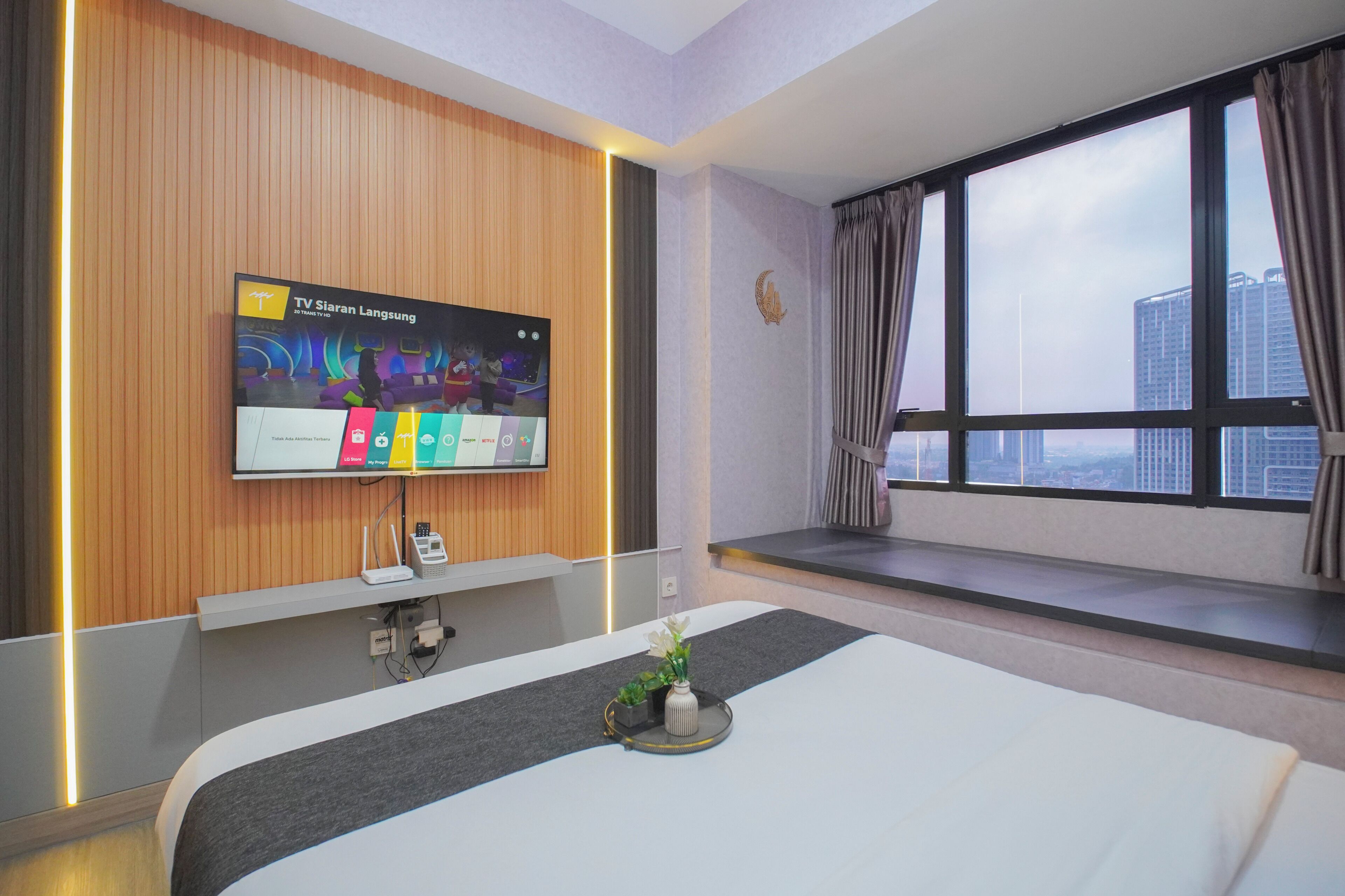 A hotel room with a large flat-screen TV mounted on the wall, a bed with a grey headboard, a small table with a vase of flowers, and a window with curtains.
