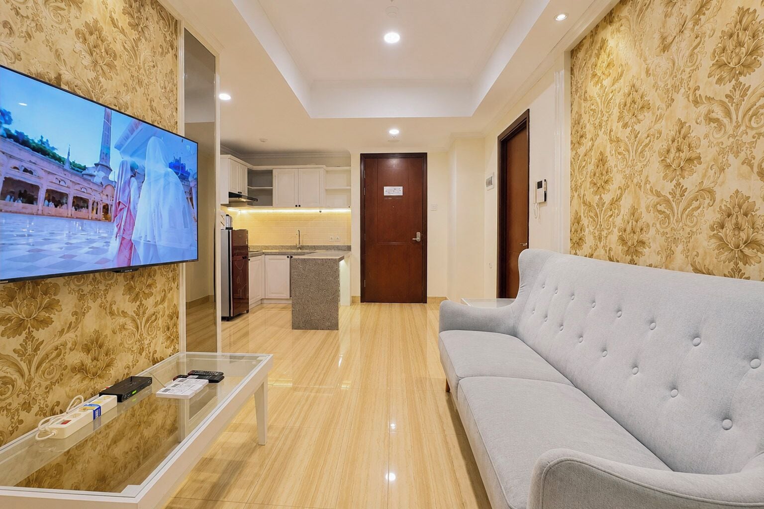 Habitación de hotel con televisión, sofá, mesa de centro y zona de cocina al fondo.