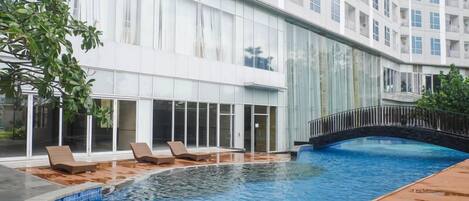 A swimming pool with lounge chairs, a wooden deck, and a building with large windows and balconies.