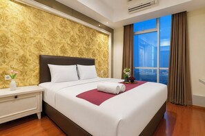 A hotel room with a large bed, a nightstand, a vase with flowers, and a view of the city through a window. - Chic And Classic 2Br At Apartment Grand Sungkono Lagoon (Surabaya)