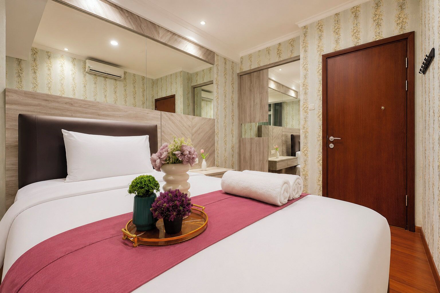 Habitación de hotel con una cama grande, cabecera de madera, un espejo, una puerta de madera y una planta pequeña sobre la cama.