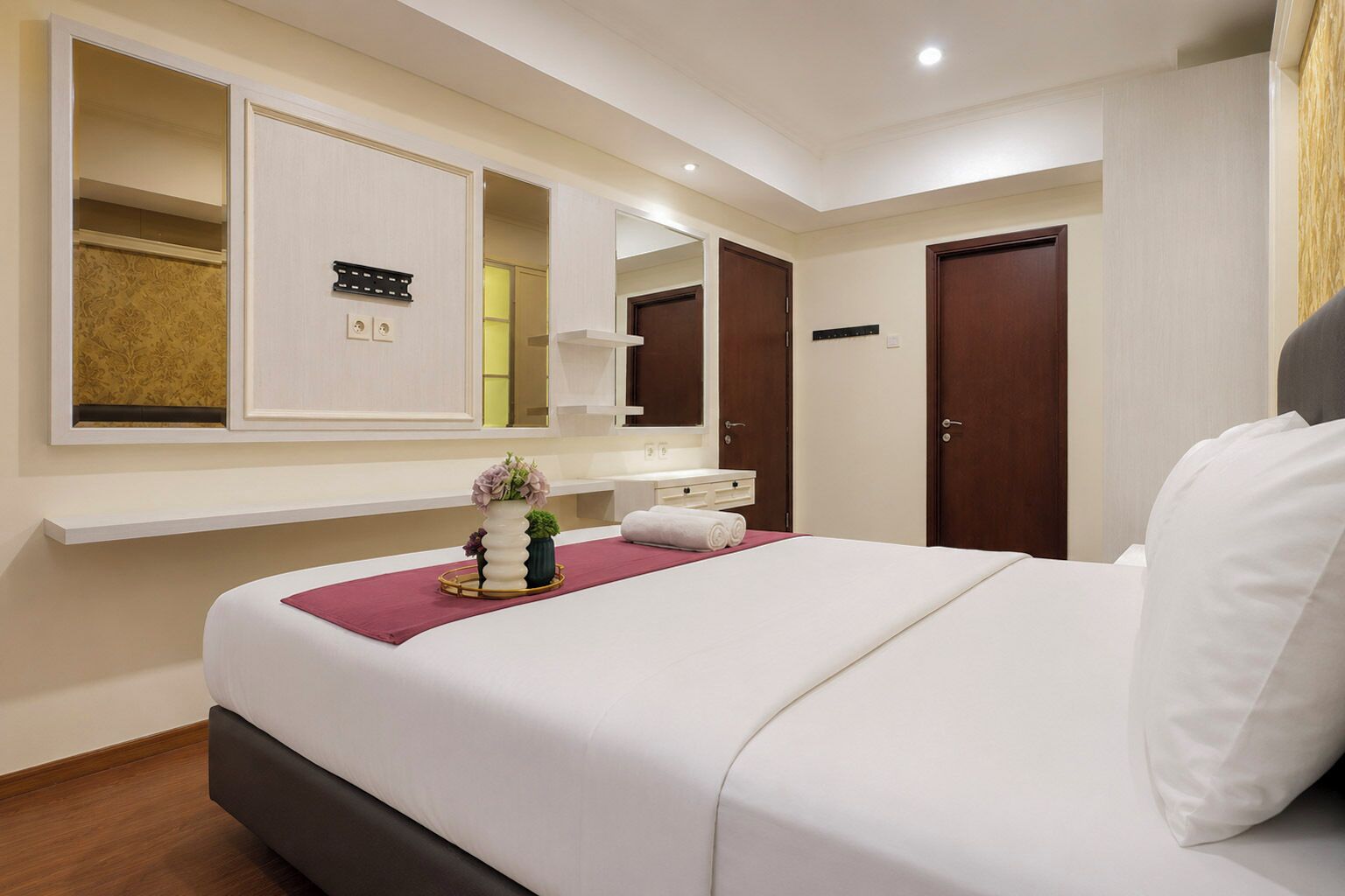 A hotel room with a bed, a vanity with a mirror, a small plant, and a wall-mounted control panel.