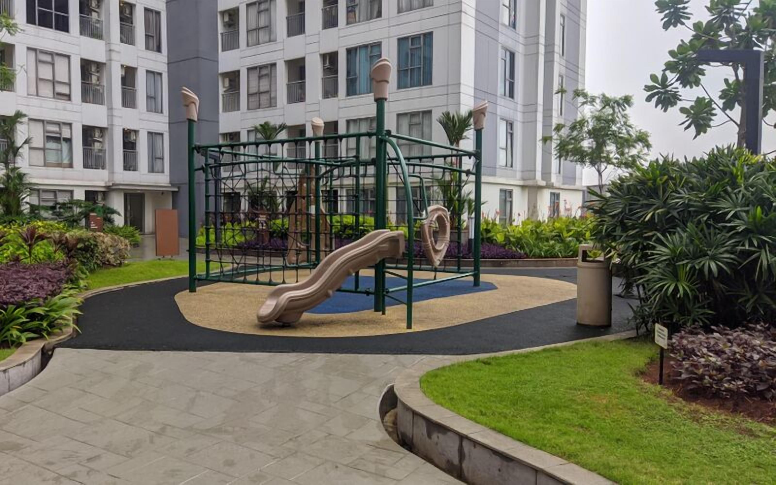 Un parque infantil con tobogán y estructuras para escalar, rodeado de un jardín ajardinado y un edificio al fondo.