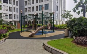 Un parque infantil con tobogán y estructuras para escalar, rodeado de un jardín ajardinado y un edificio al fondo.
