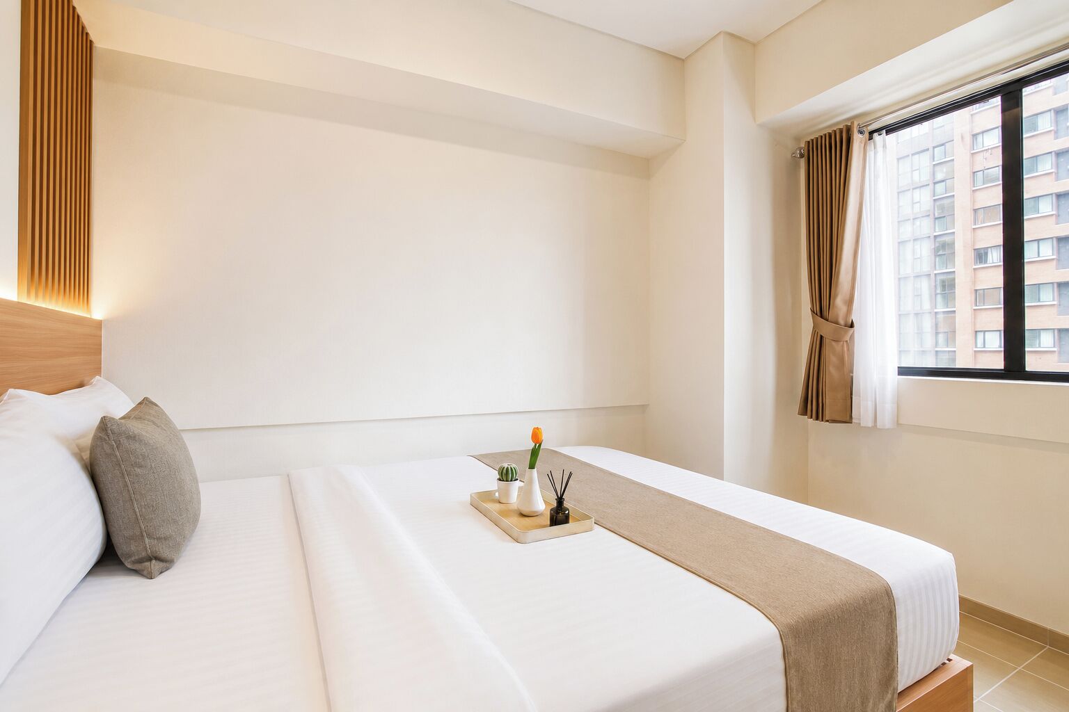 Una habitación de hotel con una cama, almohadas, una mesita de noche con un florero y una planta, y una ventana con cortinas.