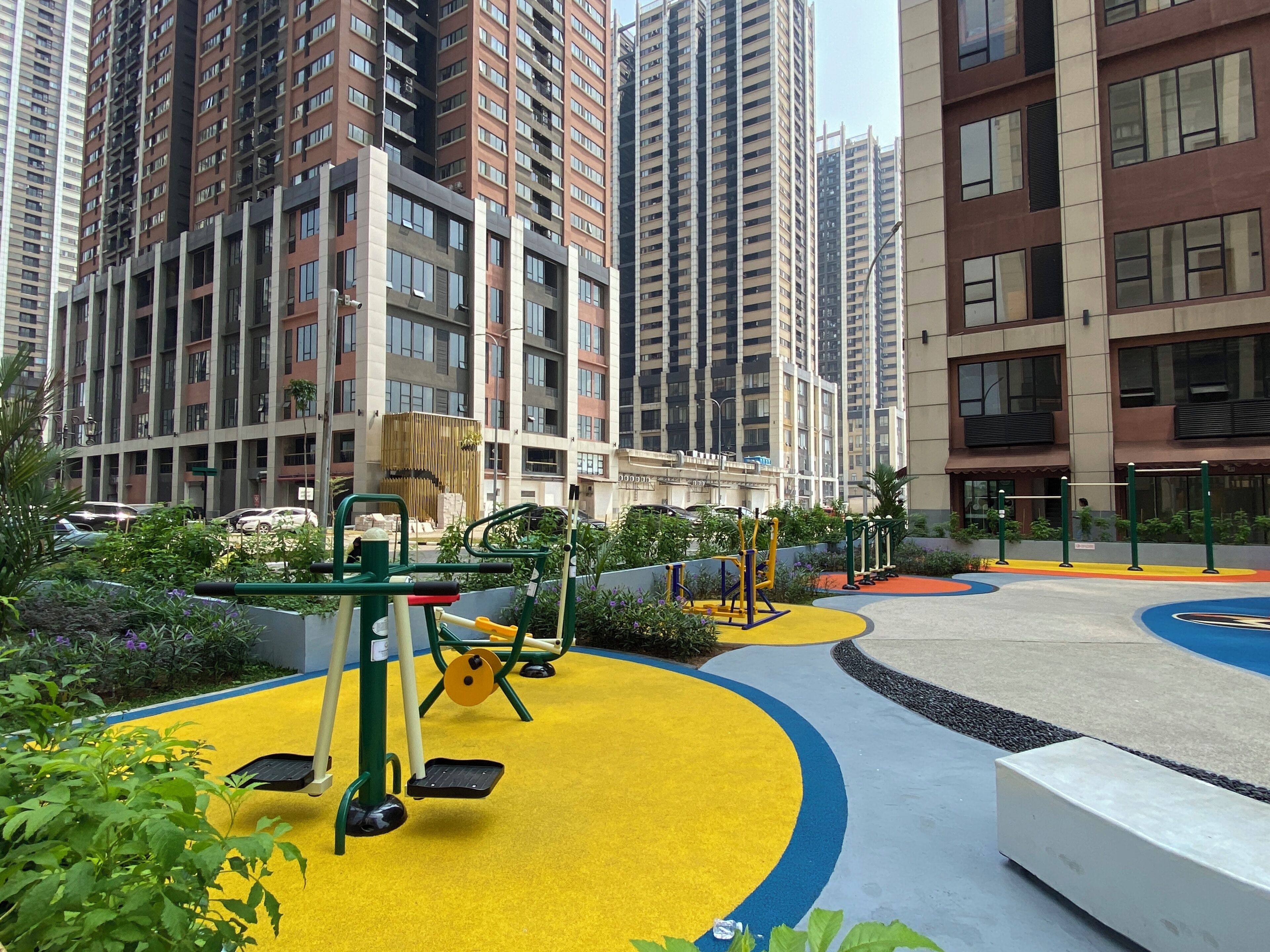 A playground with exercise equipment surrounded by greenery and buildings in the background.