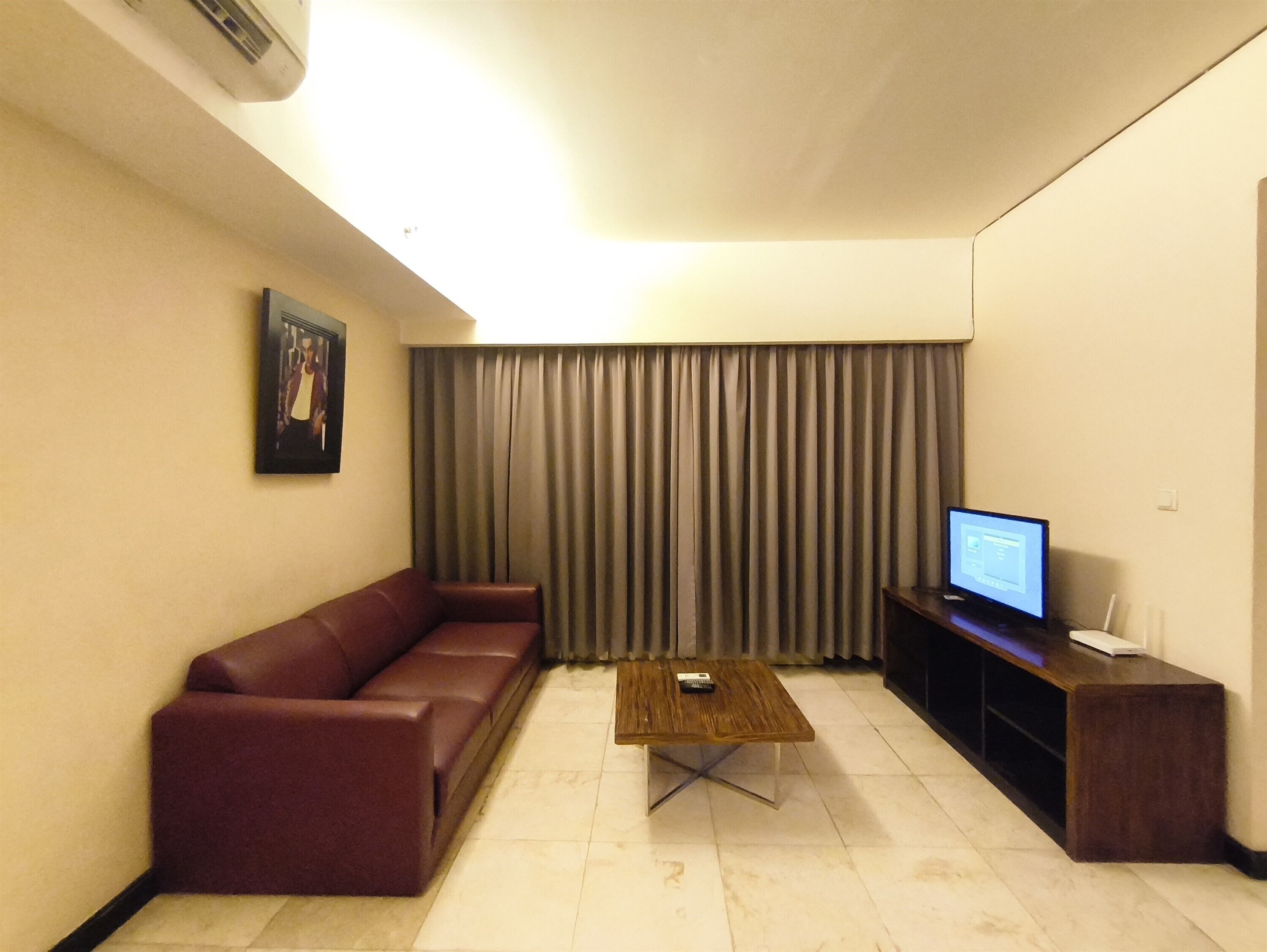 Una sala de estar con un sofá de cuero marrón, una mesa de centro de madera, un televisor sobre un mueble bajo y un ventanal con cortinas.