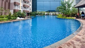 A swimming pool with a wooden deck surrounding it, a modern building in the background, and a person sitting by the pool. - Great Value 2Br At Apartment Podomoro City Deli Medan (Medan)