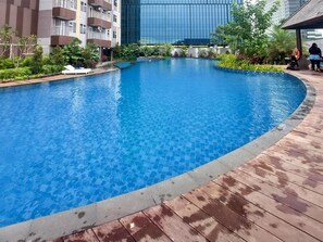 A swimming pool with a wooden deck surrounding it, a modern building in the background, and a person sitting by the pool. - Great Value 2Br At Apartment Podomoro City Deli Medan (Medan)