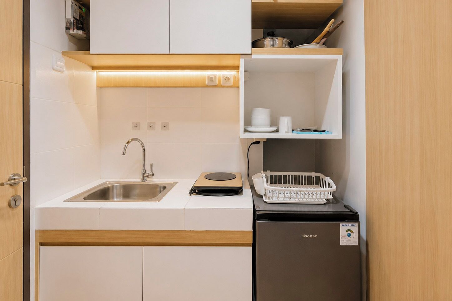 Une petite cuisine équipée avec un évier en acier inoxydable, un petit réfrigérateur et une étagère contenant des ustensiles de cuisine.