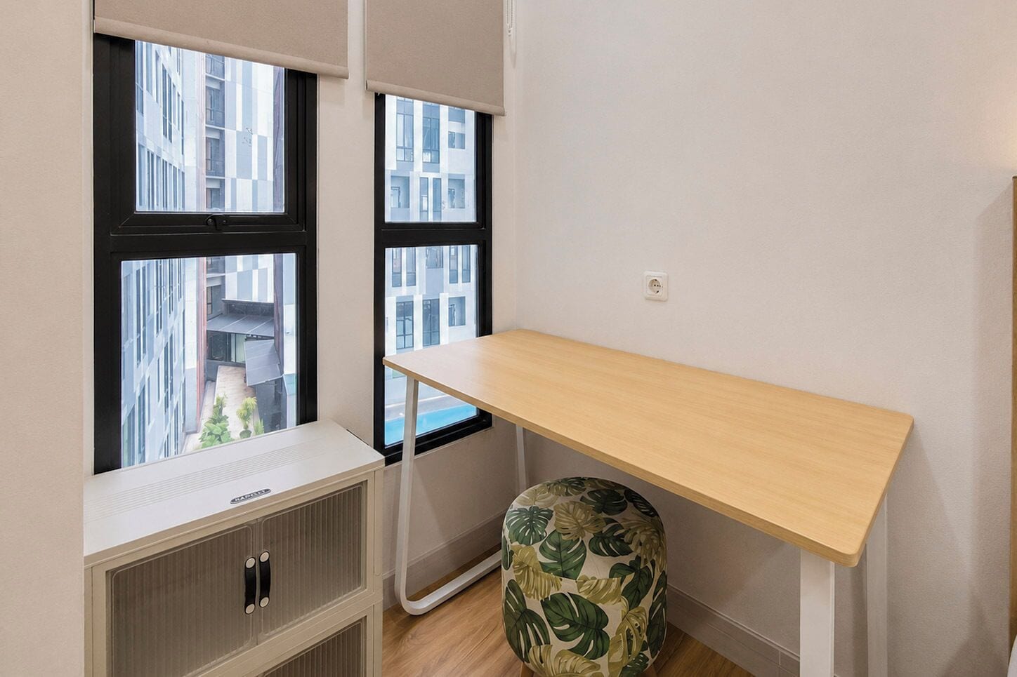 Une petite pièce avec un bureau en bois, un radiateur et une fenêtre donnant sur des immeubles.